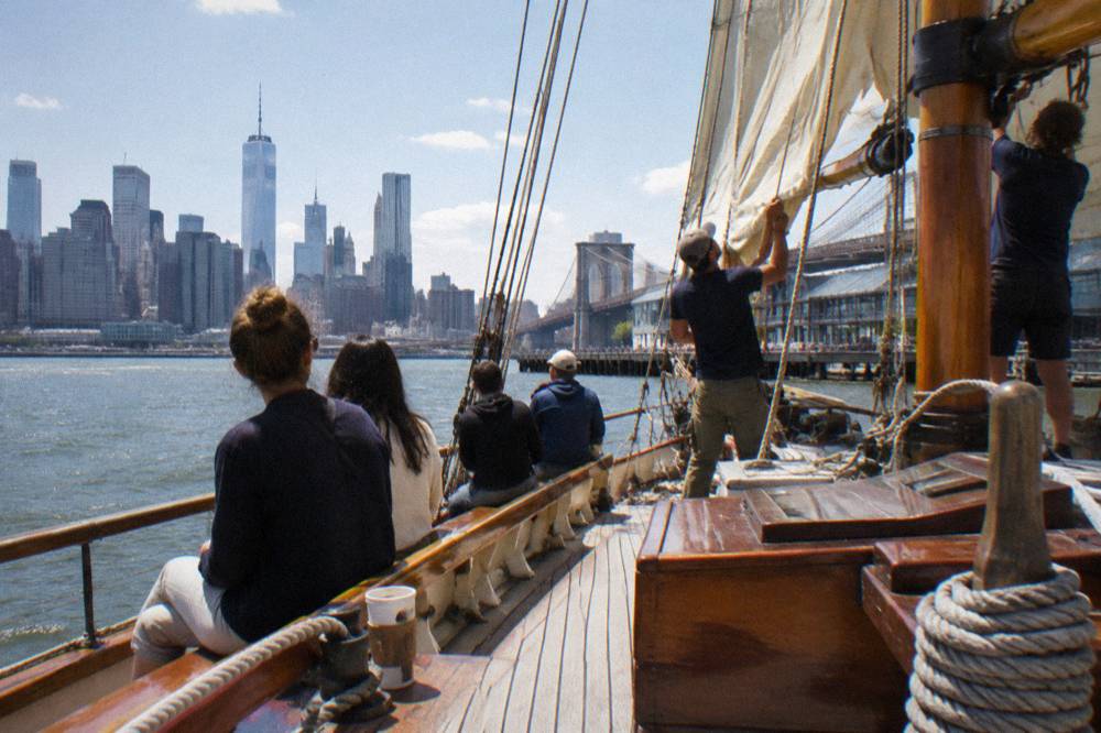 Passengers seated along the deck of a wooden schooner as crew raise the mainsail