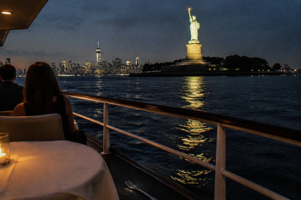 A dinner cruise boat passing the lit Statue of Liberty at night seen from the water