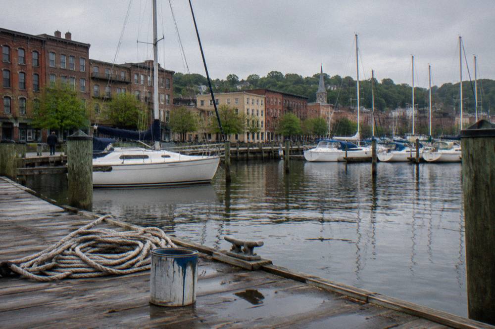 Small marina basin with masts and wooden docks beside a river town street