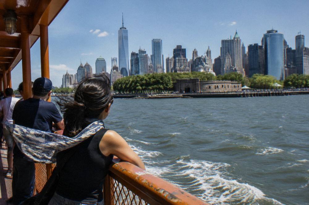 Passengers leaning on the rail of a ferry with the lower Manhattan skyline across the water
