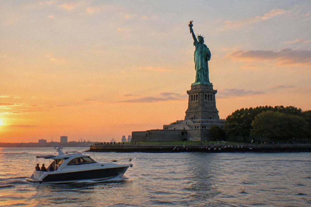 A yacht passing the Statue of Liberty in warm evening light with the harbor calm around it