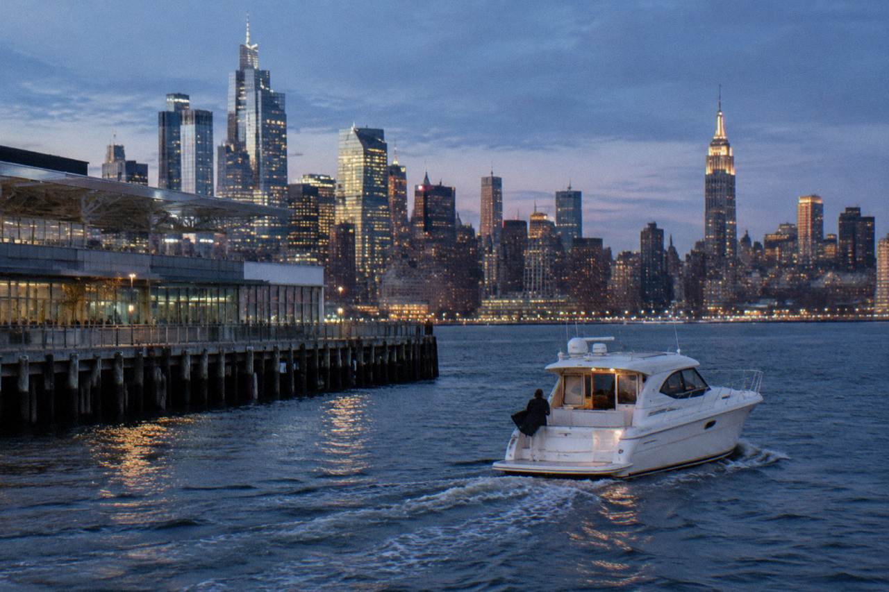 A small yacht underway on the Hudson River at dusk with the Manhattan skyline glowing behind it