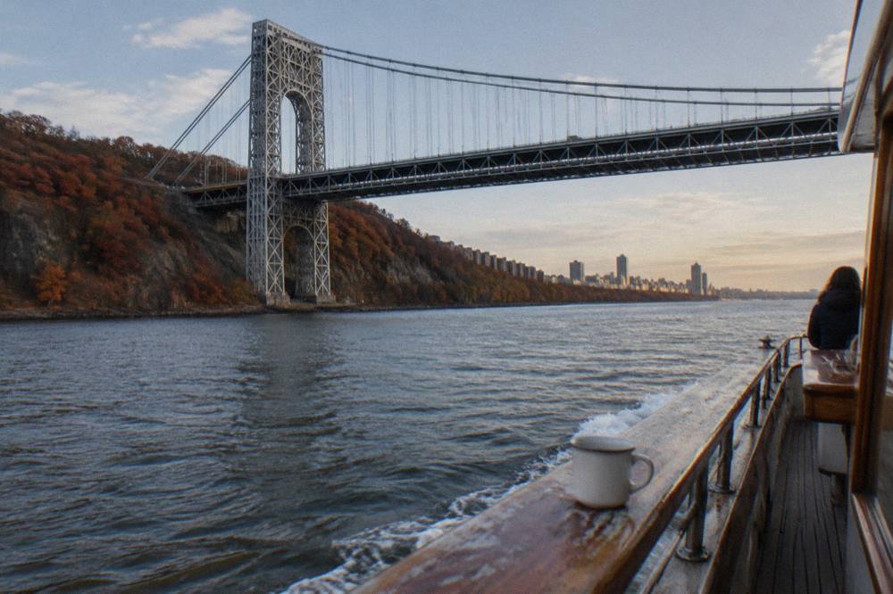A wooden yacht cruising north on the Hudson River past wooded cliffs in autumn color