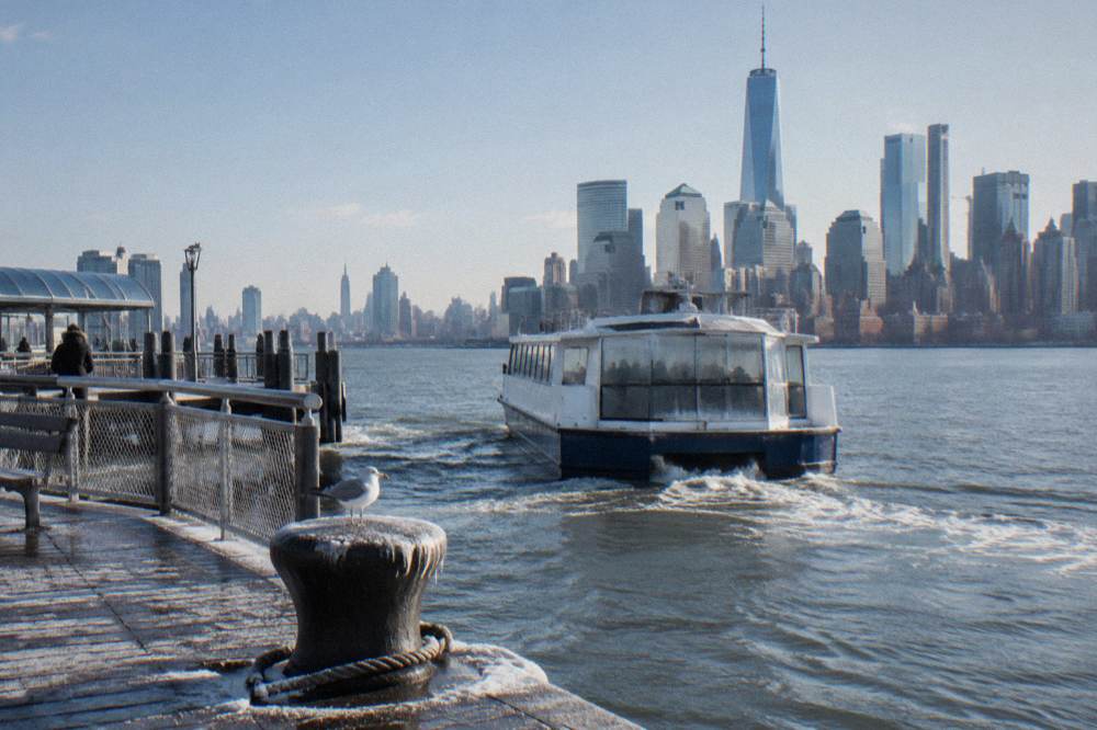 A covered sightseeing boat moving across calm harbor water toward Manhattan towers in winter light