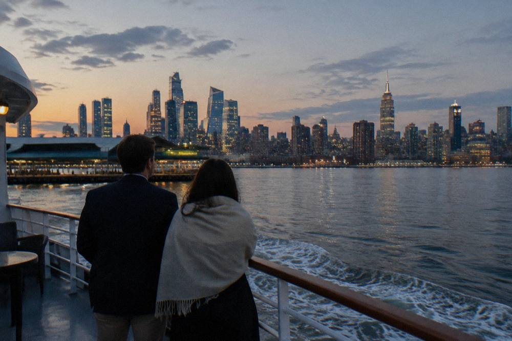 A couple in smart casual evening attire on the open deck of a yacht at dusk with the Manhattan skyline behind