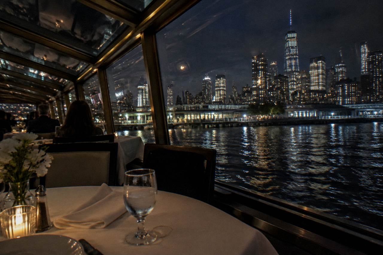 An elegant dining salon on a yacht with set tables, white linens and city lights outside the windows