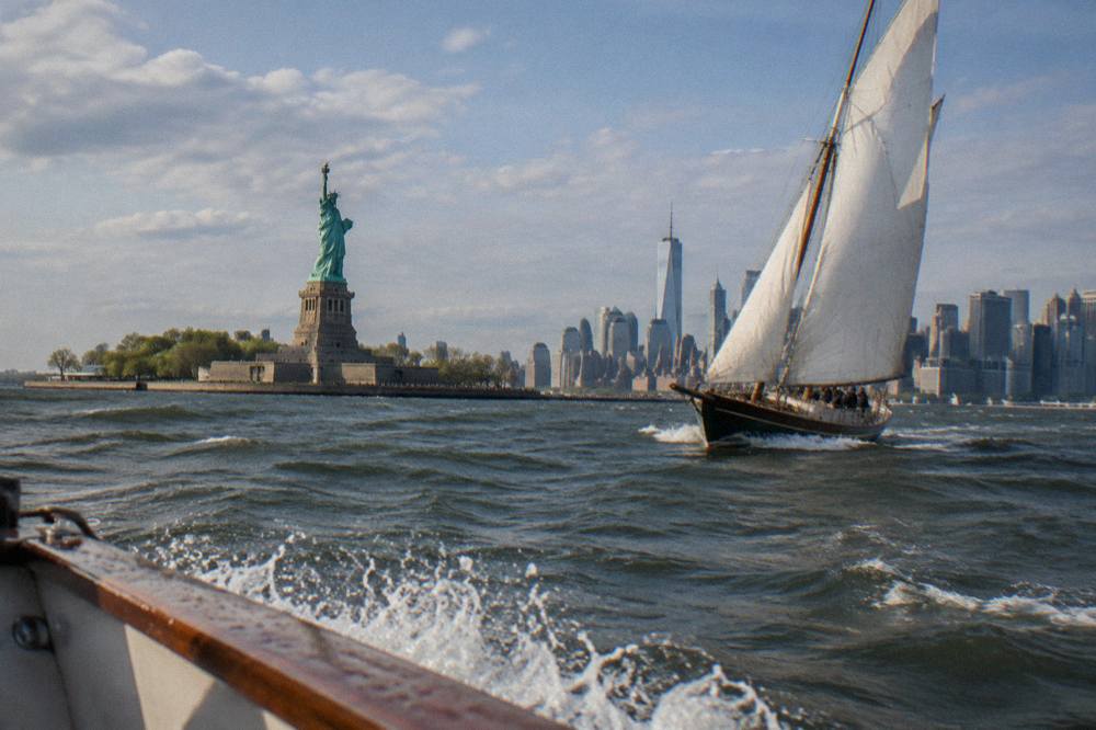 A schooner under sail on the harbor with the Statue of Liberty and the Lower Manhattan skyline behind it