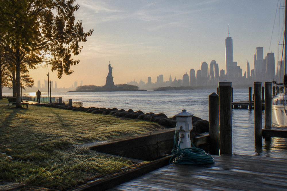 The Statue of Liberty seen across the water from a shoreline park with the skyline rising behind