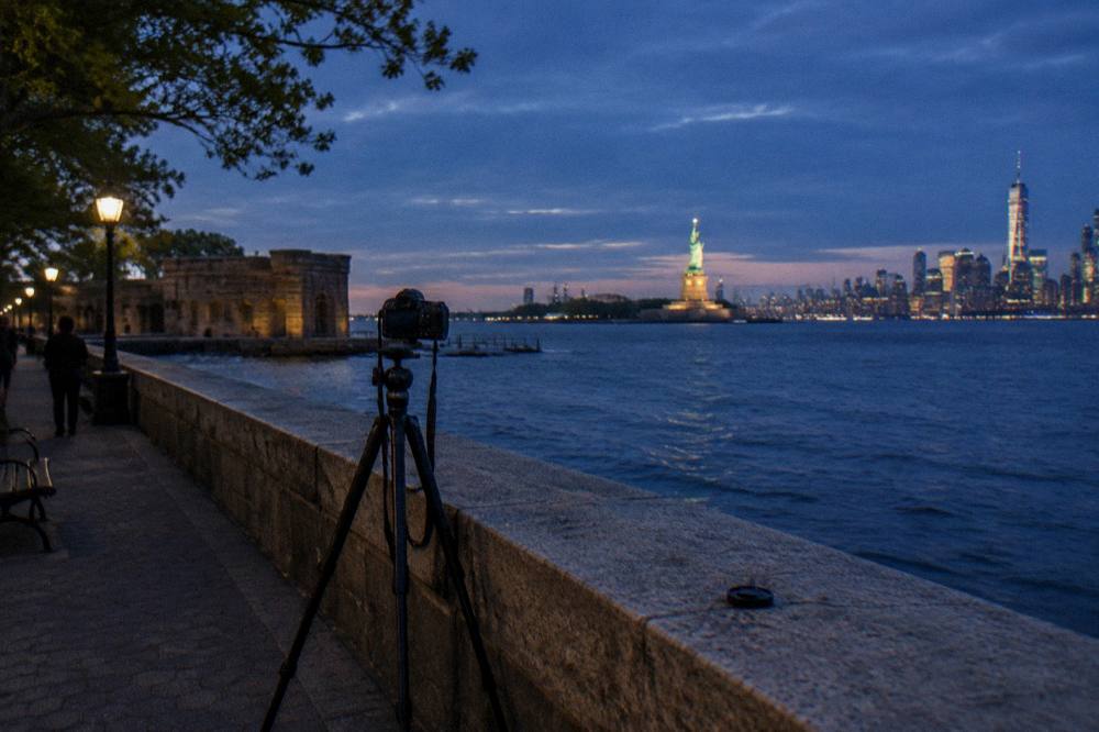 A tripod camera set up on a waterfront promenade aimed at the Statue of Liberty across the harbor at dusk