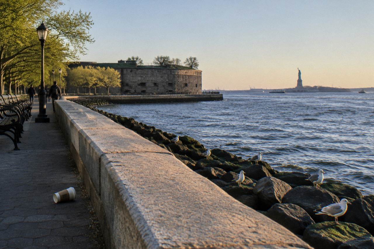 The Statue of Liberty seen across open harbor water from a Manhattan shoreline park