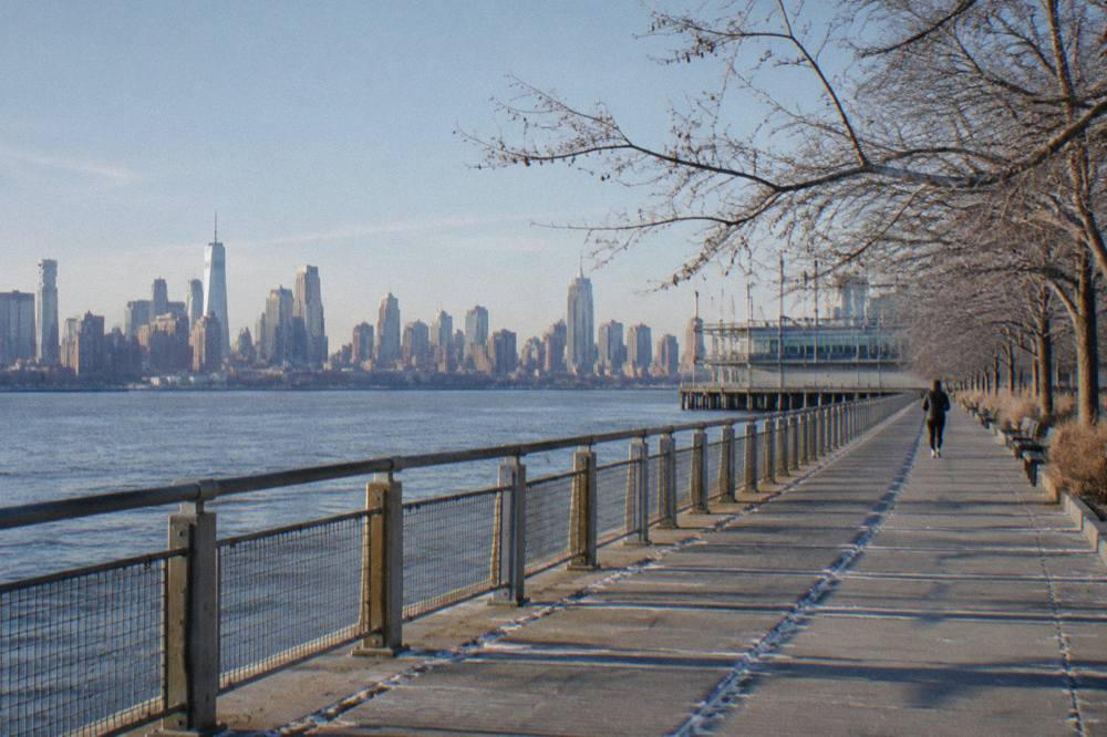 Concrete promenade along the Hudson River with benches, trees and open water beyond the railing.