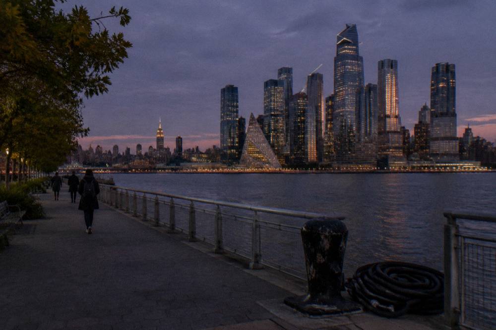 City skyline at dusk seen from a waterfront promenade with the river in the foreground.