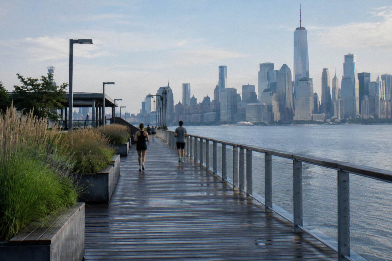 Wide view of the Hudson River waterfront with a park promenade, piers and the Manhattan skyline across the water.