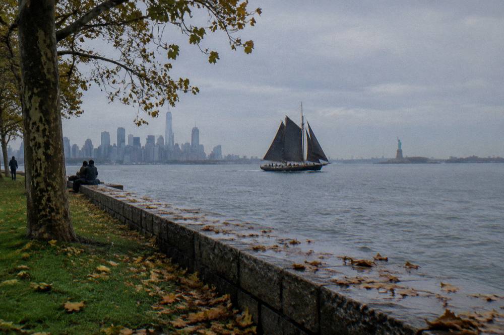 Schooner with dark sails crossing the harbor with the Statue of Liberty in the distance