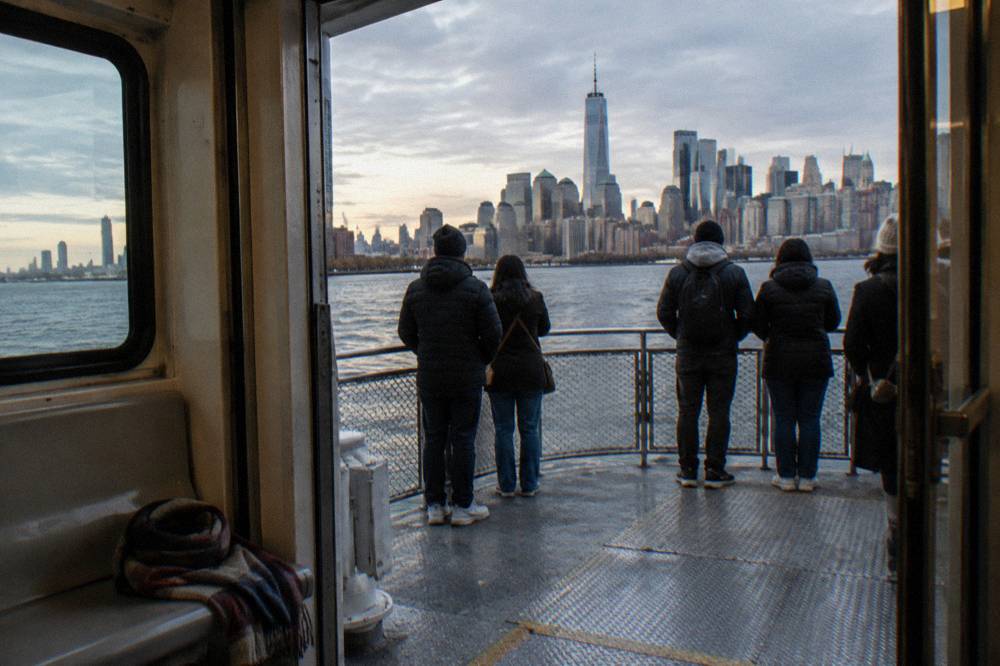 Passengers on an open deck of a sightseeing boat looking toward Lower Manhattan towers