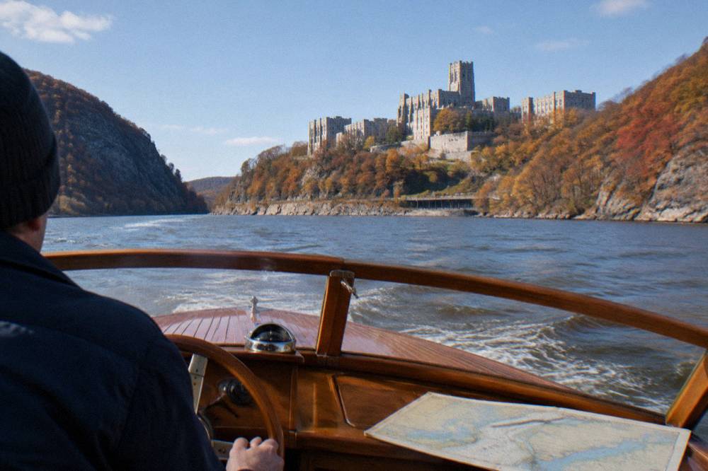 A wooden yacht motoring north on the Hudson River with autumn cliffs on both shores
