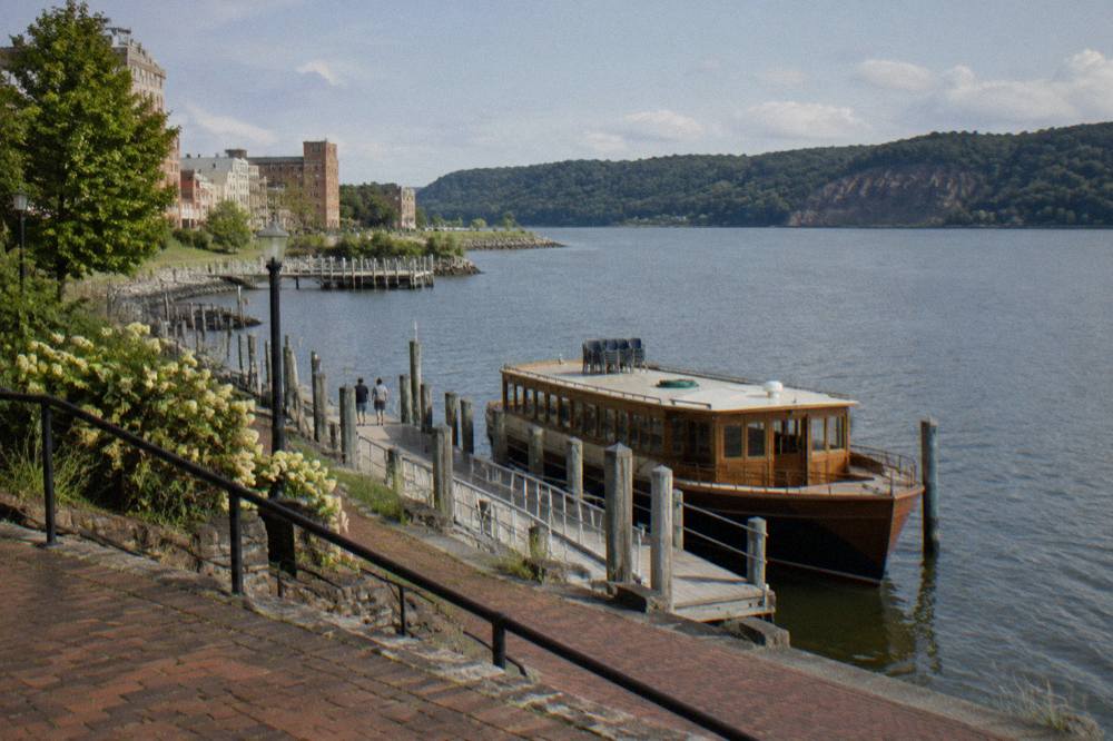 A wooden sightseeing boat docked at a river landing with wooded hills behind it