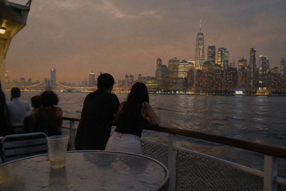 Night cruise boat deck with passengers looking toward brightly lit skyscrapers across dark water