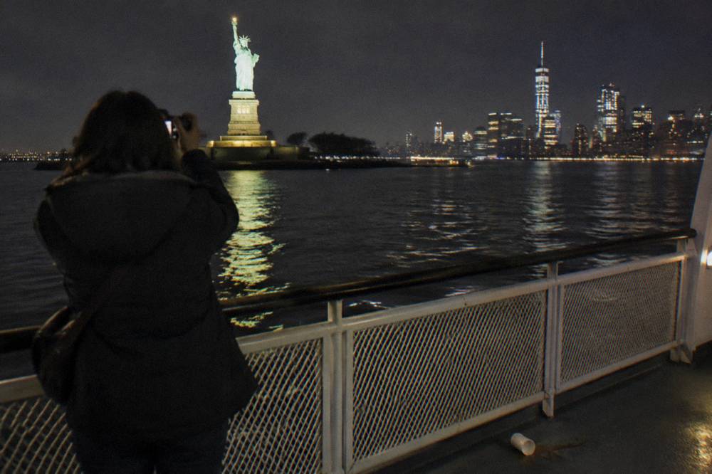 Passenger at a boat rail photographing glowing city towers across the harbor at night