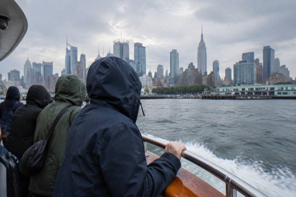 Passengers in rain jackets holding railings on a fast boat with city towers behind