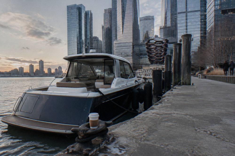 A small luxury boat moored at a Hudson River pier with glass towers behind it