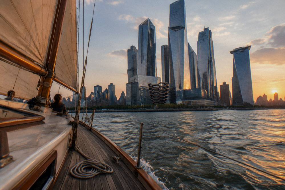 A schooner under sail on the Hudson River at sunset with city towers along the shore