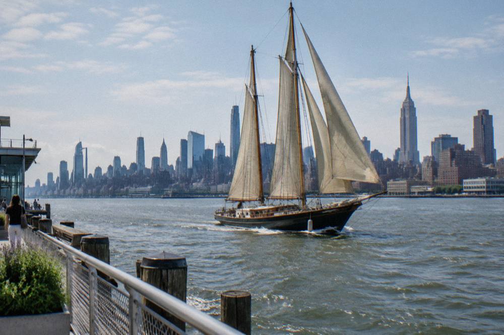 A classic schooner with tan sails crossing New York Harbor toward the Statue of Liberty