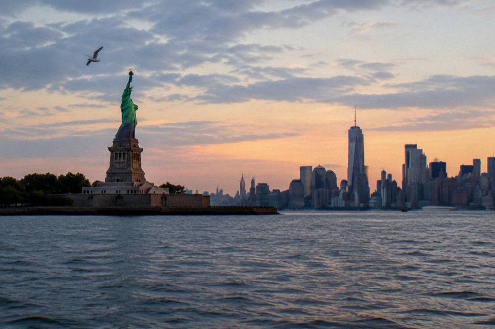 A wide view of New York Harbor at sunset with the statue and the skyline in the same frame